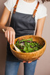 © Dina - Woman in a gray apron mixing green salad in eco wooden bamboo bowl in the kitchen. Healthy eating and nutrition, eco conscious lifestyle.