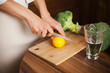 © Dina - Woman slicing a lemon on cutting board and glass of water and veggies on table - healthy lifestyle. Cutting lemon while cooking at home.