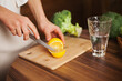 © Dina - Woman slicing a lemon on cutting board and glass of water and veggies on table - healthy lifestyle. Cutting lemon while cooking at home.