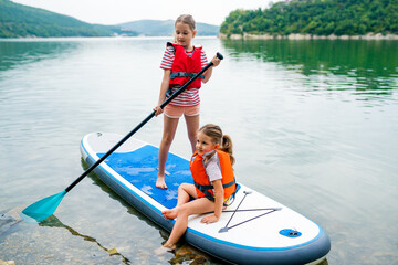 Girls padding on stand up paddle boarding on lake district. Children in swim life vest learning paddleboarding on SUP board. Family active leisure and local getaway with kids concept