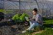 © ADDICTIVE STOCK - Farmer browsing laptop near greenhouse and wheelbarrow