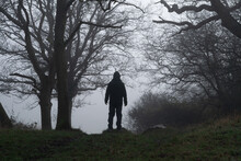 A Hooded Figure, Back To Camera Standing On The Edge Of A A Forest. On A Foggy Winters Day.