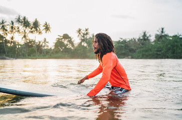 Wall Mural - Black long-haired teen man floating on long surfboard, waiting for a wave ready for surfing with palm grove litted sunset rays. Extreme water sports and traveling to exotic countries concept.