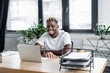 © LIGHTFIELD STUDIOS - african american man with vitiligo smiling near laptop, coffee to go and documents in office.