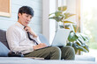 Asian thoughtful young handsome professional successful male businessman employee in formal business outfit sitting on cozy sofa couch thinking ideas working with laptop computer in living room