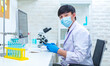 © Bangkok Click Studio - Asian professional male scientist in white lab coat and safety surgery face mask sitting typing keyboard computer analyzing experiment data information result on working desk in hospital laboratory