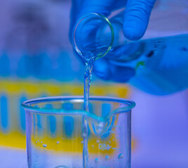 Wall Mural - Closeup studio shot of blue solution reagent liquid sample pouring from Erlenmeyer flask into glass beaker by scientist hand wearing safety rubber gloves in front of blurred test tube rack background