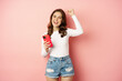 Enthusiastic brunette girl winning on mobile phone, holding smartphone and rejoicing, scream in joy, achieve app goal, pink background