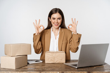 Woman small business owner, sitting at table with laptop, packing boxes, processing clients orders, showing okay, ok sign and smiling, posing against white background