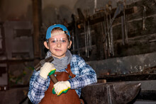 A Little Boy In A Leather Veil With A Hammer In His Hands Stands In A Blacksmith's Workshop Next To An Anvil, Is Going To Forge Iron