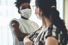 Smiling Young People Wearing Face Masks Greeting Merrily By Bumping Elbows At Workplace . Teenage Man And Woman In Facial Covers Protected From COVID-19 Coronavirus Showing Friendship And Happiness .