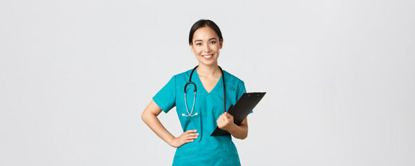 Wall Mural - Healthcare workers, preventing virus, quarantine campaign concept. Smiling pleasant asian female physician, doctor during examination wearing scrubs and holding clipboard, white background