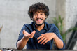 © Vadim Pastuh - Portrait of happy young Indian guy sitting at desk and gesturing. Smiling curly ethnic man in casual shirt involved virtual meeting. Hispanic male student taking a part in educational conference