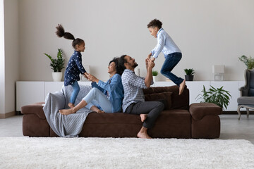 Wall Mural - Happy active little sibling kids jumping high on home couch, holding millennial parents hands for help, support. Excited family couple, son, daughter playing active games on sofa, enjoying leisure