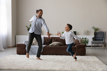 Wall Mural - Happy cheerful hipster Black dad and active preschool son kid training new dance in home living room, jumping, running, laughing in carpeted floor. Young father and child enjoying funny playtime