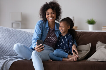 Wall Mural - Happy Black mom and cheerful kid taking selfie on smartphone, making video call, relaxing on home couch, enjoying leisure together. Young mother and little daughter using mobile phone, laughing