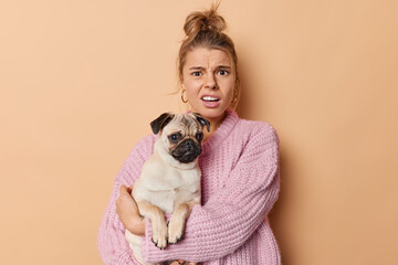 Indignant displeased young woman frowns face and looks discontent at camera hears bad news from vet about pet disease holds pedigree pug dog poses against beige background being fond of animals