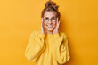 © WHstudio Leushin N - Happy young woman keeps hands on face smiles broadly shows white teeth wears round glasses for sight casual jumper isolated over yellow background. Human face expressions and emotions concept