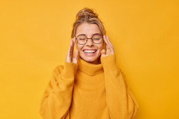 Wall Mural - Cheerful young Caucasian woman keeps eyes closed smiles toothily wears casual jumper keeps hands on rim of spectacles has good mood isolated over vivid yellow background. Happy emotions concept