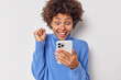 © WHstudio Leushin N - Positive optimistic young woman clenches fistt looks with excitement at smartphone screen reacts on amazing news wears blue turtleneck isolated over white background finds out she won lottery