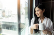 © Nutlegal - Asian beautiful young woman drinking coffee Looking through window Morning coffee is her daily routine Happy young lady hold cup of tea or hot coffee Beauty girl drinking coffee at home or hotel room