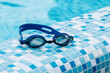 © elenakirey - Blue professional swimming goggles with water drops on yellow lenses on a blue and white tiles of pool floor.