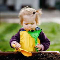 Wall Mural - Cute adorable toddler girl playing with sand and shovel on spring day. Baby child wearing yellow boots and mud rain puddle pants. Happy girl planting vegetables in spring.