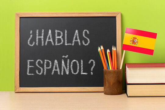A chalkboard with the question hablas espanol? do you speak Spanish? written in Spanish, a pot with pencils and the flag of Spain, on a wooden desk with green background