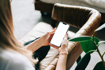 Woman holding mobile phone with white screen mock up, resting on a sofa in living room at home.	