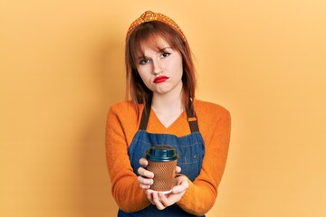 Wall Mural - Redhead young woman wearing waitress apron holding take away cup of coffee depressed and worry for distress, crying angry and afraid. sad expression.