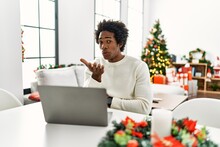 Young African American Man Using Laptop Sitting On The Table By Christmas Tree Looking At The Camera Blowing A Kiss With Hand On Air Being Lovely And Sexy. Love Expression.