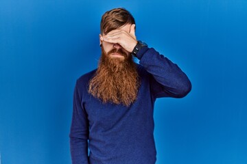 Poster - Redhead man with long beard wearing casual blue sweater over blue background covering eyes with hand, looking serious and sad. sightless, hiding and rejection concept