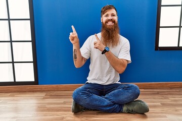 Sticker - Redhead man with long beard sitting on the floor at empty room smiling and looking at the camera pointing with two hands and fingers to the side.