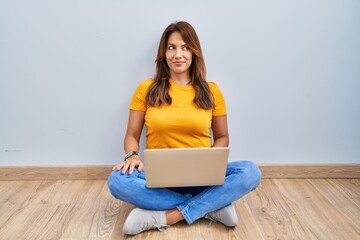 Sticker - Hispanic woman using laptop sitting on the floor at home smiling looking to the side and staring away thinking.