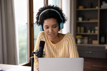 Focused pretty young African American businesswoman in wireless headphones using professional stand microphone, recording audio podcast or streaming educational video lecture looking at laptop screen.