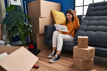 Poster - Young brunette woman sitting on the sofa at new home looking at documents annoyed and frustrated shouting with anger, yelling crazy with anger and hand raised