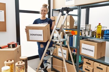 Poster - Young caucasian woman volunteer holding donations box smiling with happy face looking and pointing to the side with thumb up.