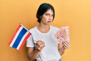 Sticker - Young caucasian woman holding thailand flag and baht banknotes clueless and confused expression. doubt concept.