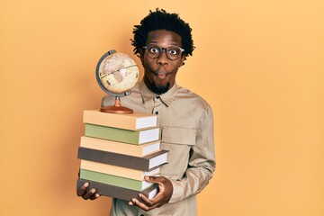 Canvas Print - Young african american man holding a pile of books and vintage world ball making fish face with mouth and squinting eyes, crazy and comical.