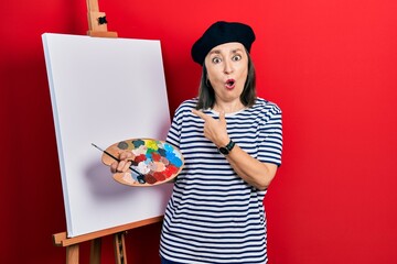 Wall Mural - Middle age hispanic woman standing drawing with palette by painter easel stand surprised pointing with finger to the side, open mouth amazed expression.