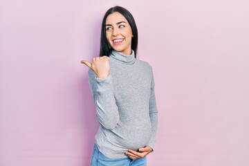 Poster - Beautiful woman with blue eyes expecting a baby, touching pregnant belly smiling with happy face looking and pointing to the side with thumb up.