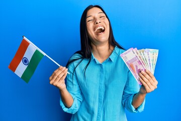 Poster - Young latin woman holding india flag and rupee banknotes celebrating crazy and amazed for success with open eyes screaming excited.