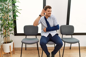 Wall Mural - Handsome young man sitting at doctor waiting room with arm injury smiling with hand over ear listening an hearing to rumor or gossip. deafness concept.