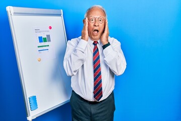 Wall Mural - Senior man with grey hair standing by business blackboard afraid and shocked, surprise and amazed expression with hands on face