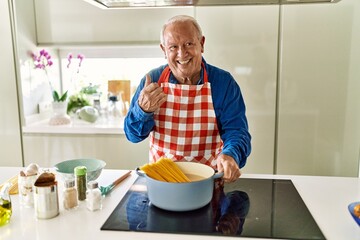 Wall Mural - Senior man with grey hair cooking spaghetti at home kitchen pointing thumb up to the side smiling happy with open mouth