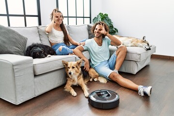 Poster - Young hispanic couple relaxing at home by vacuum robot smiling happy doing ok sign with hand on eye looking through fingers