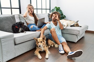 Poster - Young hispanic couple with dogs relaxing at home doing ok gesture shocked with surprised face, eye looking through fingers. unbelieving expression.