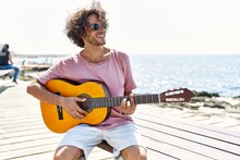 Young Hispanic Man Playing Classical Guitar Sitting On  Bench At The Beach.