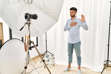 Sticker - Arab young man posing as model at photography studio swearing with hand on chest and open palm, making a loyalty promise oath