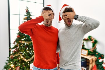 Poster - Young gay couple standing by christmas tree wearing hat peeking in shock covering face and eyes with hand, looking through fingers with embarrassed expression.